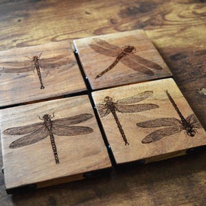 May include: Four square wooden coasters with a dragonfly design etched into the wood. The coasters are arranged in a square, showcasing the detailed insect illustrations. The wood has a warm, natural tone.