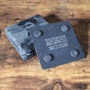 May include: A stack of square slate coasters with a black and white image of a person. One coaster shows the text "AXERAVEN MADE IN ENGLAND" and has four small, round, gray feet.