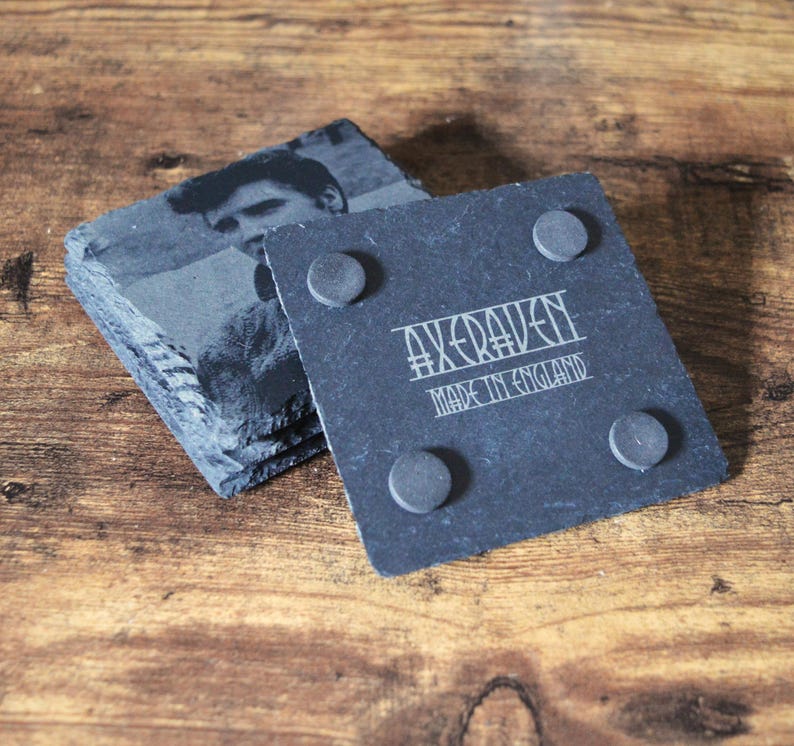 May include: A stack of square slate coasters with a black and white image of a person. One coaster shows the text "AXERAVEN MADE IN ENGLAND" and has four small, round, gray feet.