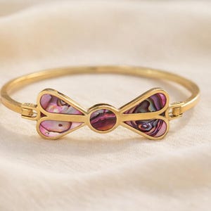 Puede incluir: Pulsera dorada con un centro en forma de lazo. El lazo está incrustado con concha de abulón iridiscente multicolor, que muestra tonos rosas, morados y azules. La pulsera tiene un diseño articulado y un cierre seguro.