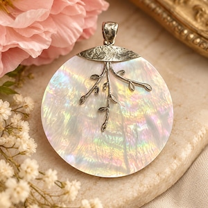 May include: A circular pendant featuring a polished, iridescent mother-of-pearl disc. A textured, silver-toned metal bail sits at the top, extending into a delicate, branch-like metal overlay with small leaves centered on the shell surface.