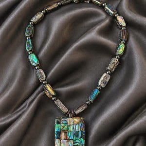 Puede incluir: Un collar con un colgante rectangular y una serie de cuentas rectangulares iridiscentes. El colgante y las cuentas muestran un mosaico de colores, incluyendo azules, verdes y morados. El collar está sobre un fondo de satén gris.