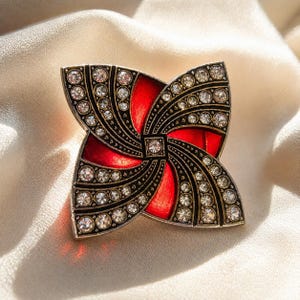 May include: A decorative pin with a four-petal design. The pin features a vibrant red enamel center, framed by black filigree and encrusted with sparkling clear crystals. The pin is set against a soft, cream-colored fabric.