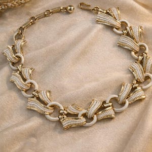 Pode incluir: Um colar vintage dourado com detalhes em esmalte branco e um design decorativo. O colar apresenta uma série de elementos interconectados, criando uma peça ousada e elegante. O fecho é visível, e o colar é exibido em uma superfície macia de tom neutro.