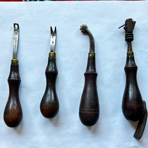May include: Four antique leather working tools with wooden handles. The tools have different metal tips for stitching, cutting, and shaping leather.