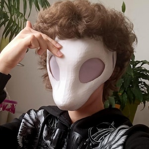 Puede incluir: Una máscara blanca y lisa que cubre todo el rostro con grandes aberturas oculares ovaladas de color claro. La máscara tiene una forma cónica similar a un hocico y se lleva con una sudadera negra con cremallera y estampado gráfico.