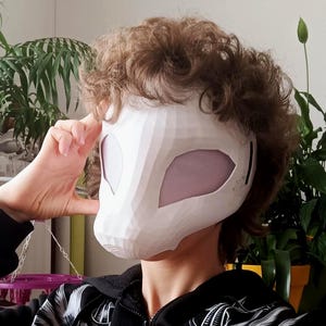 Puede incluir: Una máscara geométrica blanca de estilo low-poly con grandes aberturas oculares en forma de almendra cubiertas por una malla fina de color claro. La máscara tiene una superficie lisa y facetada que cubre la parte superior del rostro.