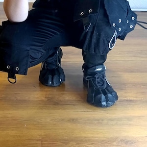 Puede incluir: Pantalones cargo negros con m&uacute;ltiples bolsillos, ojales met&aacute;licos plateados y herrajes en forma de D. Los pantalones se combinan con zapatillas negras con cordones que presentan un dise&ntilde;o de superposici&oacute;n segmentada sobre el empeine.