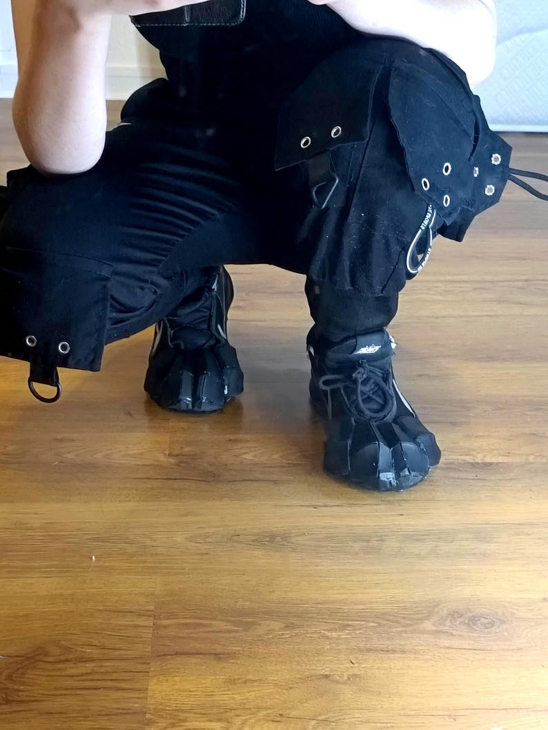 Puede incluir: Pantalones cargo negros con m&uacute;ltiples bolsillos, ojales met&aacute;licos plateados y herrajes en forma de D. Los pantalones se combinan con zapatillas negras con cordones que presentan un dise&ntilde;o de superposici&oacute;n segmentada sobre el empeine.