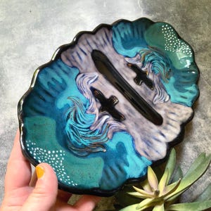May include: An oval ceramic dish with a scalloped edge, featuring a glossy glaze in shades of teal, blue, and white. The surface displays a painted landscape with two black bird silhouettes, swirling water patterns, and white dotted accents.