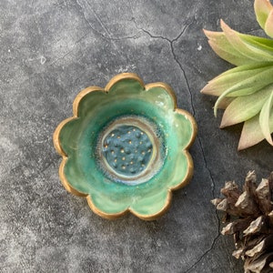 May include: A handmade, ceramic, flower-shaped dish with a teal glaze and gold trim. The center of the dish is a darker teal with gold speckles. The dish is a decorative item.
