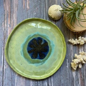 May include: A green ceramic plate with a blue and black flower design in the center. The plate has a slightly raised rim and a glossy finish.