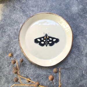 May include: A cream-colored ceramic plate with a black and white moth design in the center. The plate has a gold-colored rim. The moth has white spots on its wings. The plate is on a gray surface.
