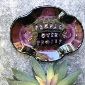 May include: A ceramic ashtray with a black and purple glaze. The words "PEOPLE OVER PROFIT" are embossed in the center. The ashtray has a unique, organic shape and is decorated with gold accents.