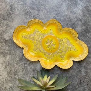 May include: A cloud-shaped ceramic tray with a yellow and gold color scheme. The tray features a floral design in the center and along the edges. The tray is approximately 10 inches long and 7 inches wide.