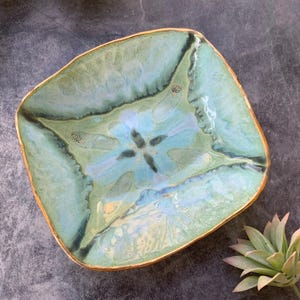 May include: A square ceramic dish with a light blue and green glaze. The dish has a gold rim and a floral design in the center.