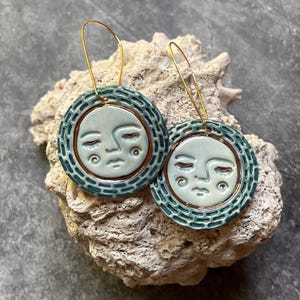 May include: A pair of round ceramic earrings featuring a light blue face with closed eyes and a textured dark teal border. Each earring has a gold-toned wire hook and a metallic copper-colored rim separating the face from the patterned edge.