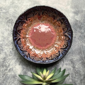 May include: A handmade ceramic bowl with a floral pattern. The bowl features a dark purple rim, a reddish-pink center, and orange and brown floral designs. The bowl is a decorative piece for serving food or displaying items.