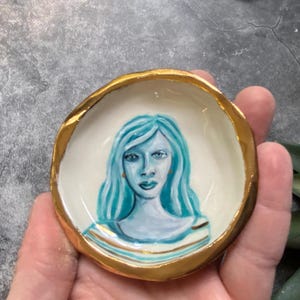 May include: A small white ceramic dish with a gold rim. The dish has a painted portrait of a woman with blue hair and blue eyes.
