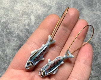 May include: A pair of fish-shaped earrings. The earrings feature a light blue and white color scheme with gold accents. The fish have dark spots and are suspended from gold-colored ear wires. The earrings are a unique accessory.