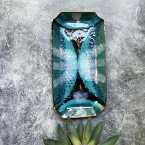 May include: A rectangular ceramic tray with beveled edges, featuring a glossy glaze in shades of teal, turquoise, and deep black. The surface displays a symmetrical, abstract pattern with a metallic gold-painted rim around the entire perimeter.