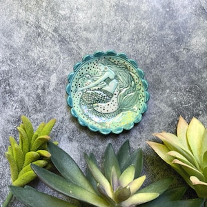 May include: A small, round, turquoise ceramic dish with a scalloped edge. The dish features a detailed illustration of a mermaid with long flowing hair and a scaled tail. The mermaid is surrounded by small black dots and gold accents.