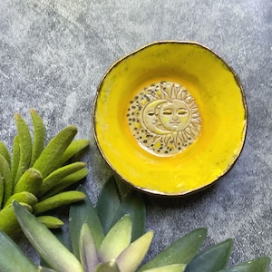 May include: A small, handmade ceramic dish with a bright yellow interior and a gold-colored rim. The center features a detailed relief of a sun and moon design. The dish is surrounded by green and gray artificial succulents.