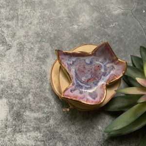 May include: A small, star-shaped ceramic dish with a purple and gold glaze. The dish is sitting on a wooden coaster.
