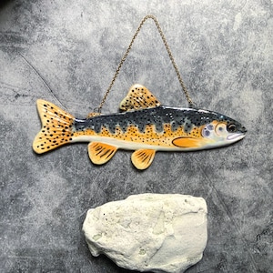 May include: A ceramic fish wall hanging with a detailed design. The fish features a yellow and black spotted tail, fins, and dorsal fin. The body is a mix of yellow, black, and white. A gold chain is attached for hanging. The fish is approximately 12 inches long.