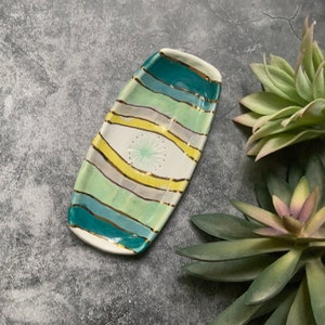 May include: A white ceramic dish with a rectangular shape and a decorative design of green, blue, and yellow stripes. The dish has a gold trim and a small, white, star-shaped design in the center.