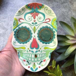 May include: A ceramic skull-shaped dish with a colorful Day of the Dead design. The dish features a white base with teal, green, red, and gold accents. The skull design includes floral and geometric patterns.