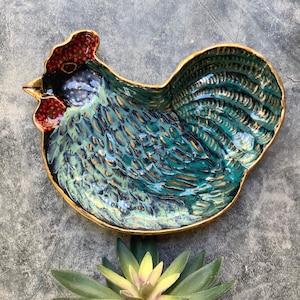 May include: A ceramic rooster-shaped dish with a teal and gold glaze. The dish has a red comb and a gold rim. The design is detailed, with textured feathers and a beak. The dish is a decorative item.