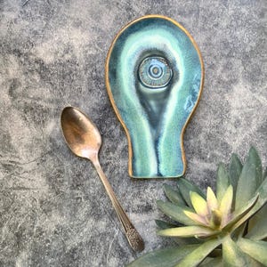 May include: A handmade ceramic spoon rest in shades of teal and blue with a gold rim. A vintage silver spoon rests beside the spoon rest. A succulent plant is in the lower right corner. The spoon rest has a unique eye-like design.
