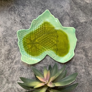 May include: A green ceramic dish shaped like a leaf with a textured surface. The dish has a green glaze with a darker green pattern.