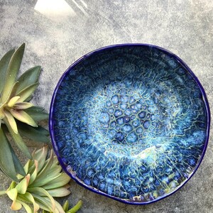 May include: A handmade ceramic bowl with a deep blue glaze. The bowl's interior features a textured, bubbly pattern in shades of blue and turquoise, with a dark blue rim. The bowl is round and appears to be sitting on a gray surface.