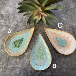 May include: Three ceramic trinket dishes in the shape of teardrops with gold trim. The dishes are decorated with a green and white paisley design and have a light blue, light pink, and white glaze.