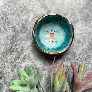 May include: A small, round ceramic trinket dish with a glossy finish. The interior features a light blue center that transitions to a deep teal rim with a metallic gold edge. The text LOVE YOU is stamped in black with a small orange heart in the center.