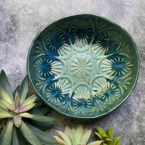 May include: A handcrafted ceramic bowl with a detailed floral pattern in shades of teal and green. The bowl's rim is a darker teal, with lighter teal and green accents in the center. The bowl is approximately 10 inches in diameter.
