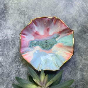 May include: A decorative ceramic bowl with a ruffled edge and a gold rim. The bowl features a blend of pink, blue, and white colors, with a textured, turquoise center. The bowl is placed on a gray surface, with a green succulent plant in the foreground.
