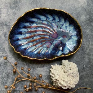 May include: A handcrafted ceramic serving dish with a unique, organic shape. The dish features a deep blue glaze with streaks of brown and purple, and a gold-colored rim. Decorative and functional, it is suitable for serving appetizers or displaying small items.