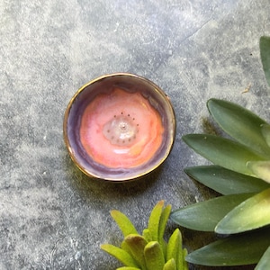May include: A small, round ceramic bowl with a gold rim. The interior features a floral design with shades of pink, orange, and purple. The bowl is likely handmade and suitable for holding small items. The background is a gray surface with green plant accents.