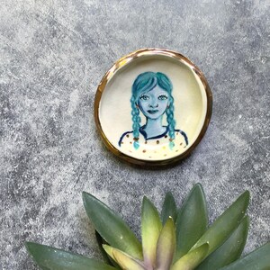 May include: A round ceramic trinket dish with a metallic gold rim. The center features a hand-painted illustration of a person with blue hair in two braids, wearing a cream-colored shirt with small brown polka dots against a white background.