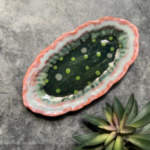 May include: An oval ceramic dish with a dark green interior speckled with light green and white dots. The rim is a mix of pink, white, and green. The dish is on a gray surface, with a succulent plant in the corner.