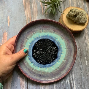 May include: A ceramic plate with a purple, blue, and green glaze. The center of the plate features a black and white design with a radial pattern.