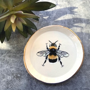 May include: A small, round, white ceramic plate with a gold rim. The plate features a detailed illustration of a bumblebee in black, yellow, and white. The plate is approximately 5 inches in diameter.
