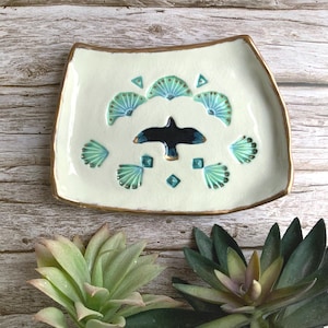 May include: A white ceramic dish with a gold rim and a black bird design. The dish is decorated with green and blue fan-shaped patterns.