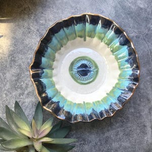 May include: A ceramic bowl with a unique design. The bowl features a white interior with a central eye-like design in shades of blue and green. The rim is ruffled and edged with a gold color, and the exterior is painted with blue and green hues.
