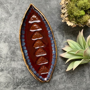 May include: A red ceramic dish with a gold rim and blue accents. The dish is decorated with a repeating pattern of stylized sunbursts.