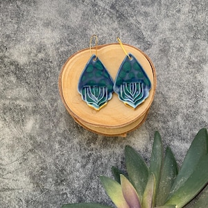 May include: A pair of teardrop-shaped ceramic earrings with a blue and green glaze and gold accents. The earrings have a floral design and are hanging from gold hoops.