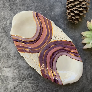 May include: A white ceramic serving dish with a unique design featuring purple and gold stripes. The dish has a wavy, organic shape and is perfect for serving snacks or appetizers.